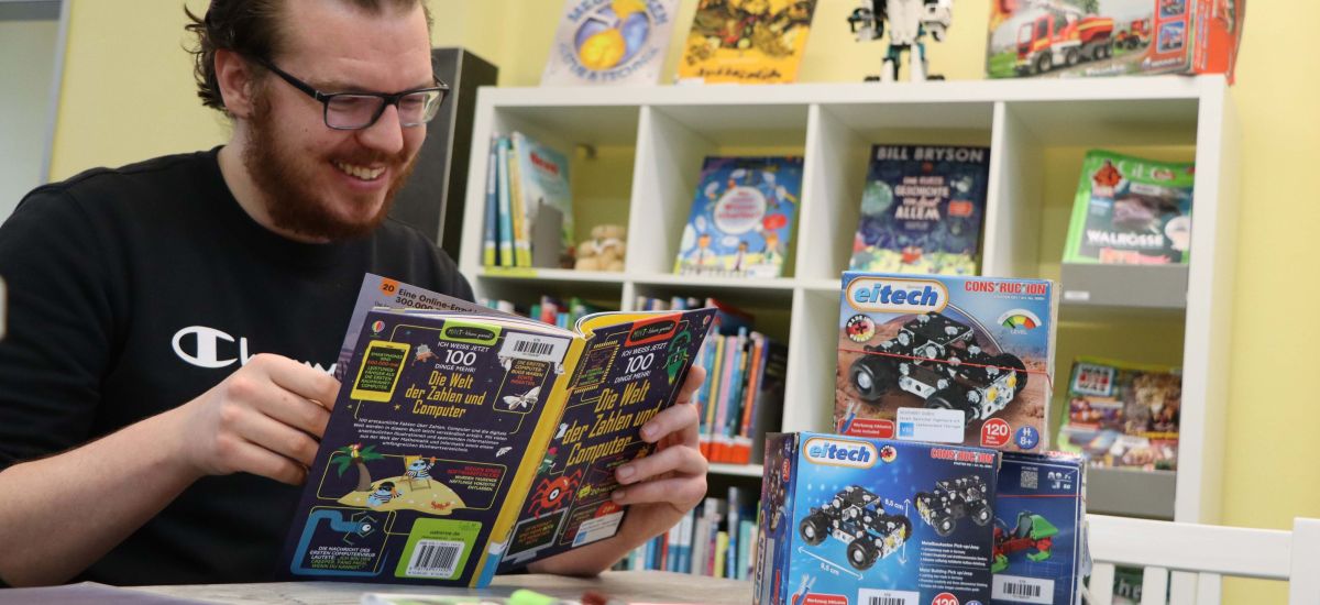 Ein Mann sitzt an einem Tisch und ließt in einem Buch. Im Hintergrund steht ein Regal mit Büchern.