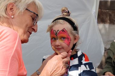 Eine Frau malt einem Kind einen roten Schmetterling ins Gesicht.
