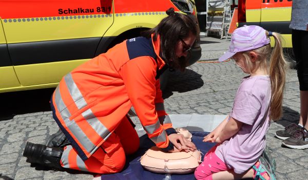 Ein Frau in oranger Uniform zeigt einem Kind die Herzdruckmassage an einer Puppe.