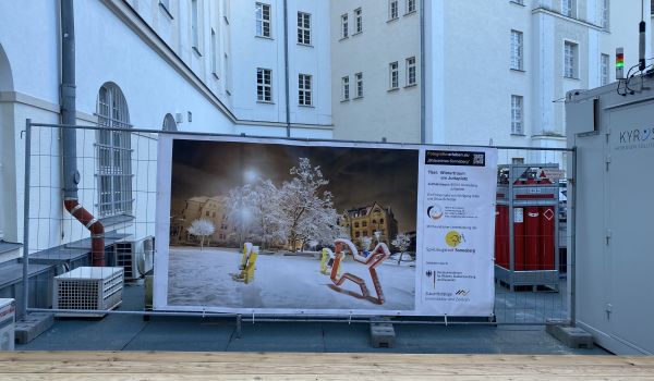 An einem Bauzaun hängt ein großes Banner. Darauf ist der Juttaplatz in Sonneberg im Winter zu sehen.