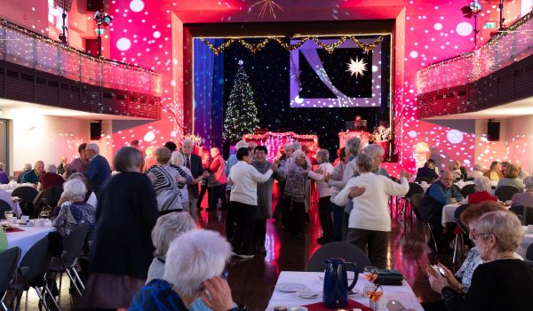Senioren tanzen in einem großen Saal. Im Hintergrund ist eine Bühne mit großem Weihnachtsbaum.