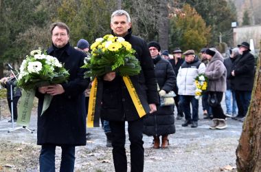 Zwei dunkel gekleidete Männer tragen Blumengebinde zu einem Gedenkstein.