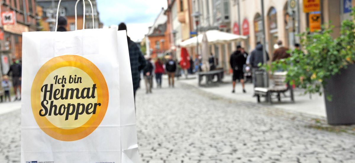 In der belebten Einkaufsstraße steht im Vordergrund eine weiße Papier-Einkaufstasche mit dem Slogan "Ich bin Heimat Shopper".