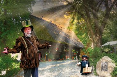 Ein als Spielmann verkleideter Mann steht vor einem alten Holzhaus.