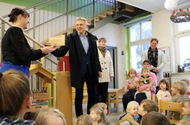 Ein Mann übergibt einen Umschlag an eine Frau in einem Kindergarten. Rundherum sitzen Mädchen und Jungen, die sich über diese Nikolaus-Überraschung freuen.