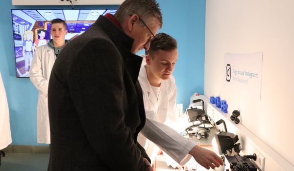 Ein Jugendlicher in weißem Kittel erklärt Experimente für Interessierte.