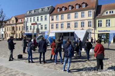 Eine Traube von Menschen steht auf einem Marktplatz vor einem geöffneten mobilen Wagen mit Informationen zum Thema Internet.