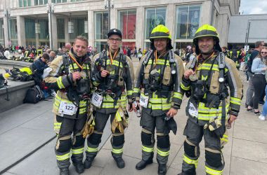 Vier Feuerwehrmänner in Schutzkleidung stehen nebeneinander auf einem großen Platz mit vielen Menschen. Sie tragen eine Medaille um den Hals.