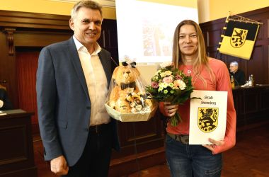 Eine Frau ält eine Urkunde und einen Blumenstrauß und bekommt vom Bürgermeister noch einen Teddy überreicht.