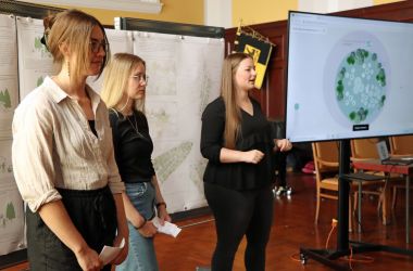 Drei junge Frauen stehen vor Plakaten. Sie halten Karteikarten in den Händen. Rechts daneben steht ein Monitor mit dem Plan einer Grünanlage darauf.