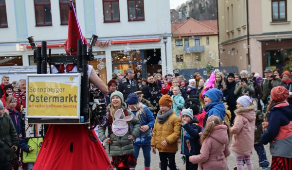 Ein rot kostümierter Mann trägt ein Schild auf dem Rücken mit der Aufschrift: Sonneberger Ostermarkt mit verkaufsoffenem Sonntag. Mit einem Gerät erzeugt er Seifenblasen. Um ihn herum versammeln sich viele Kinder.