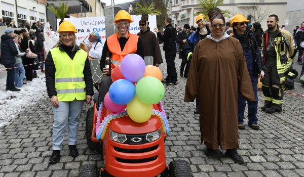 Ein geschmückter Rasentraktor. Dahinter laufen Personen, die als Bauarbeiter gekleidet sind.