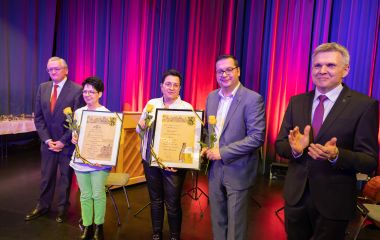 Auf einer Bühne stehen der Bürgermeister und Professor Dippold, welche ein Präsent an drei Personen überreichen.