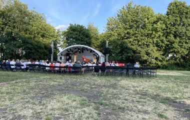 Auf einer Wiese steht eine Bühne. Davor sitzen Menschen auf Stühlen.