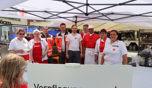 Mitarbeiter des DRK stehen an hinter einem Stand. Davor steht ein Schild mit der Aufschrift: Verpflegungsausgabe.