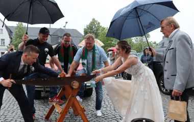 Ein Brautpaar sägt einen Baumstamm durch. Außenherum stehen Leute.