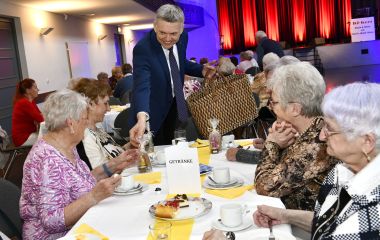 Senioren sitzen an gedeckten Kaffeetafeln in einem großen Saal. Ein Mann verteilt Geschenke.