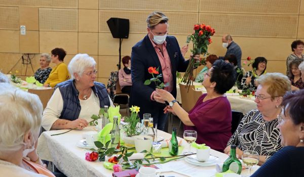 Frauen sitzen an einem Tisch. Ein Mann verteilt Rosen an sie.