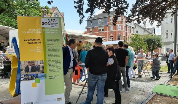 Menschen stehen neben einem Banner und unterhalten sich. Auf dem Banner steht NEC SON - Zusammen wachsen.