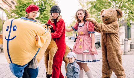 Vier Personen in Spielzeugkostümen stehen in einer Reihe und halten sich an den Schultern. Vor ihnen steht ein kleines Kind.