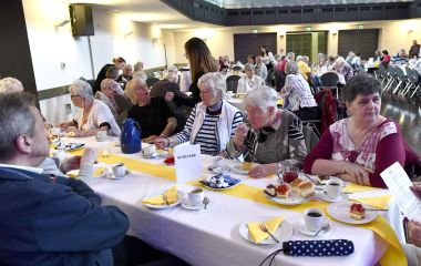 Senioren sitzen an gedeckten Kaffeetafeln in einem großen Saal.