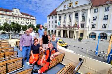 In einem Bus ohne Dach stehen und sitzen mehrere Menschen. Im Hintergrund ist das Sonneberger Rathaus zu sehen.