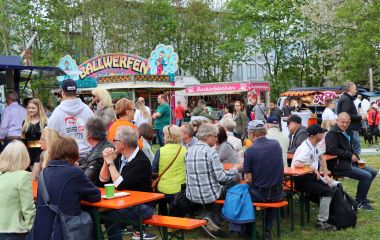Viele Menschen sitzen auf Bierzeltgarnituren auf einer Wiese. Im Hintergrund stehen verschiedene Jahrmarktattraktionen.