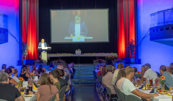 Der Bürgermeister steht auf einer Bühne und spricht. Davor sitzen Menschen.