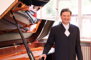 Ein schwarzgekleideter Mann steht vor einem Flügel und hält sich mit einer Hand daran fest.