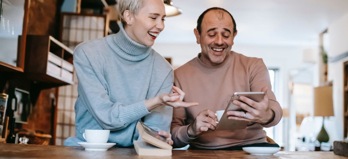 Eine junge Frau mit einem Buch und ein Herr mit einem Tablet schauen lachend auf das Tablet, Pexels.com / Gary Barnes