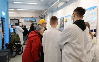 Jugendliche in weißen Kitteln erklären Experimente für Interessierte.