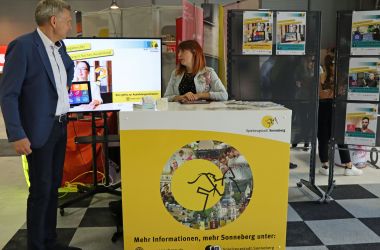 Eine Frau steht hinter einem gelben Messestand. Auf der Standvorderseite ist das Logo der Stadt Sonneberg - das Sonneberger Reiterlein. Daneben steht der Bürgermeister. Im Hintergrund steht ein Bildschirm mit der Aufschrift: Ausbildungsberufe.