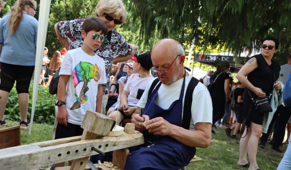 Ein Mann fertigt ein kleines Schiff aus Holz. Um ihn herum stehen Menschen.