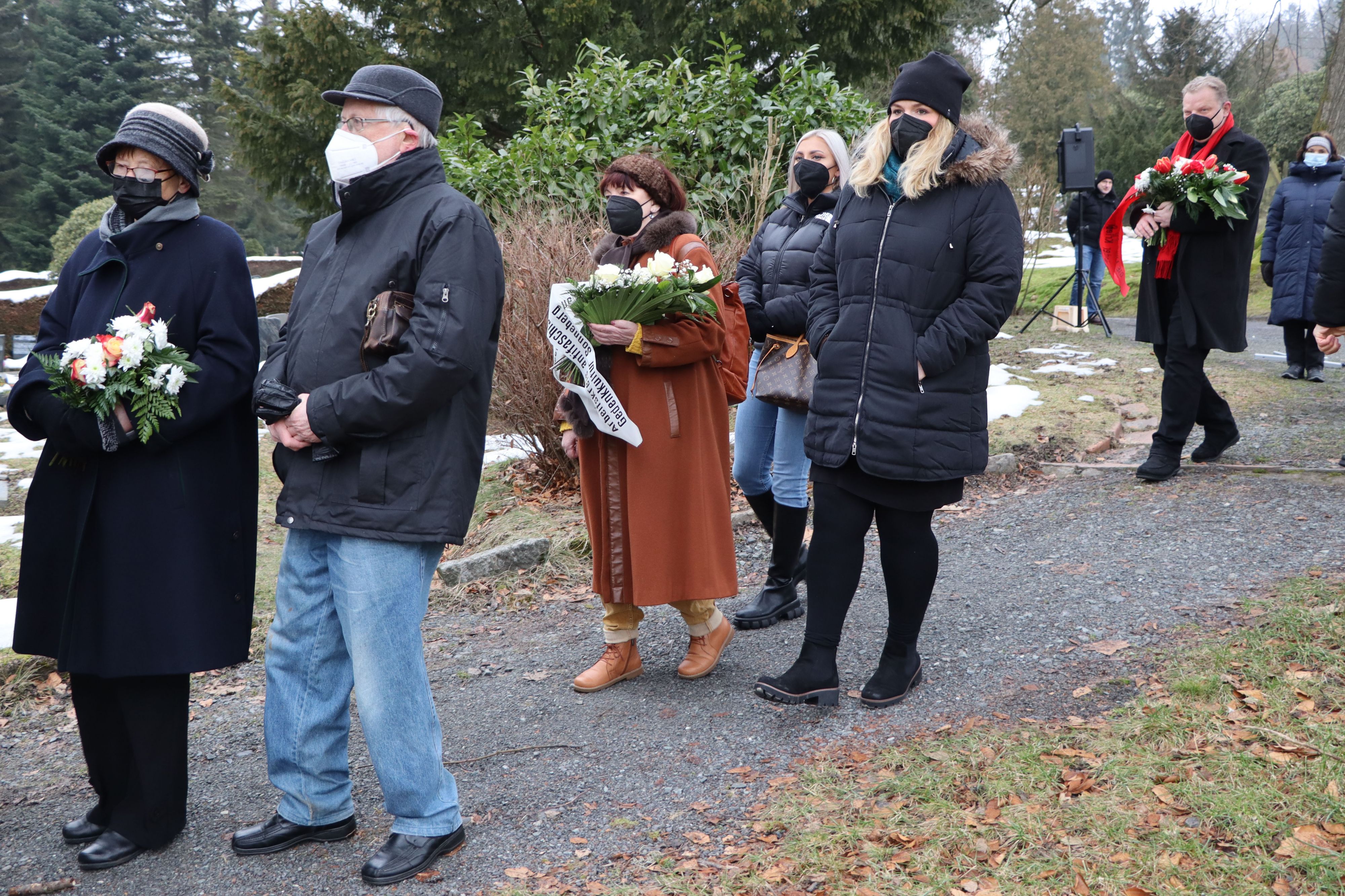 Mehrere Personen mit Blumensträußen in der Hand laufen über den Stadtfriedhof.