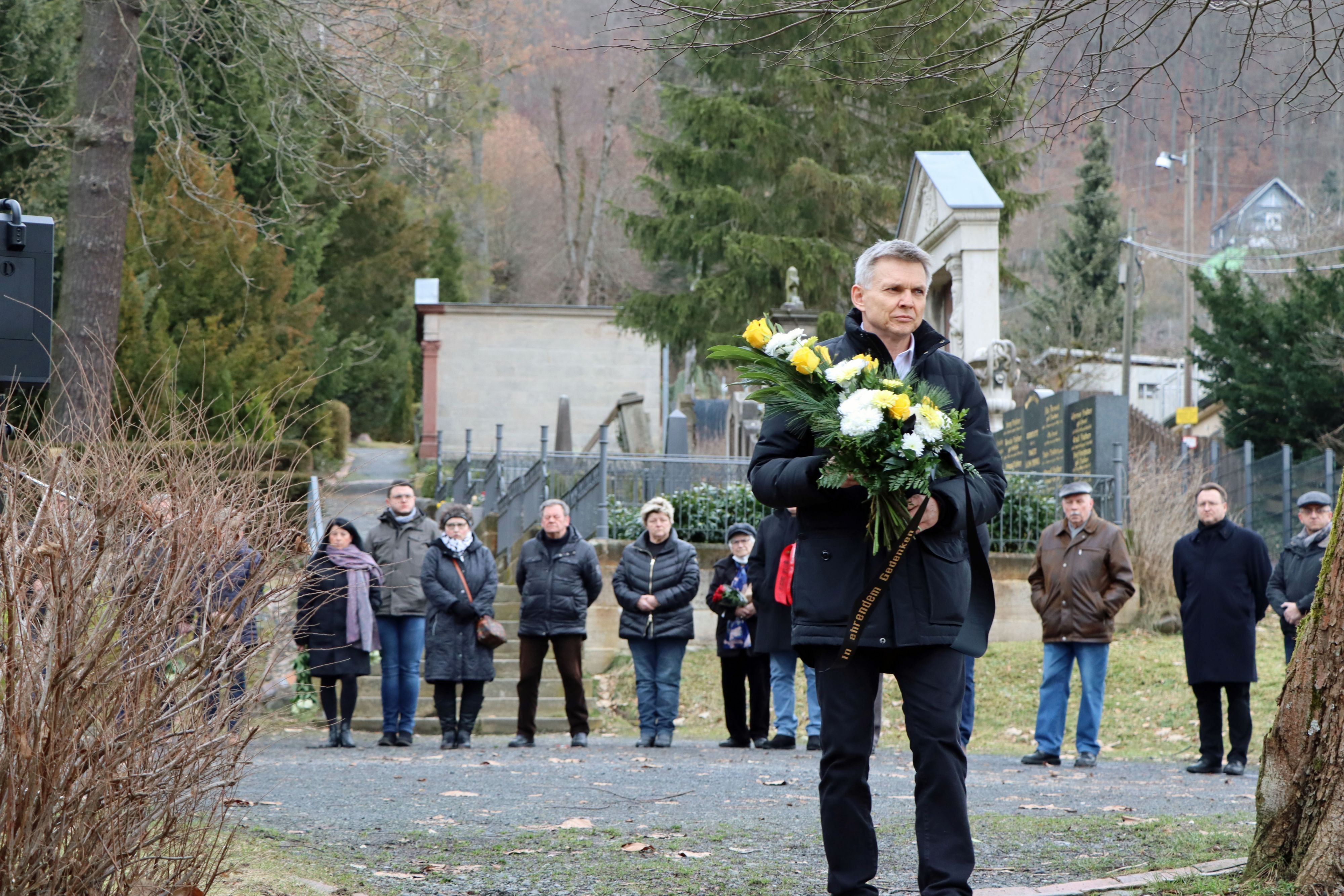 Ein dunkel gekleideter Mann steht mit einem Blumenstrauß auf dem Friedhof. Im Hindergrund stehen weitere Personen.