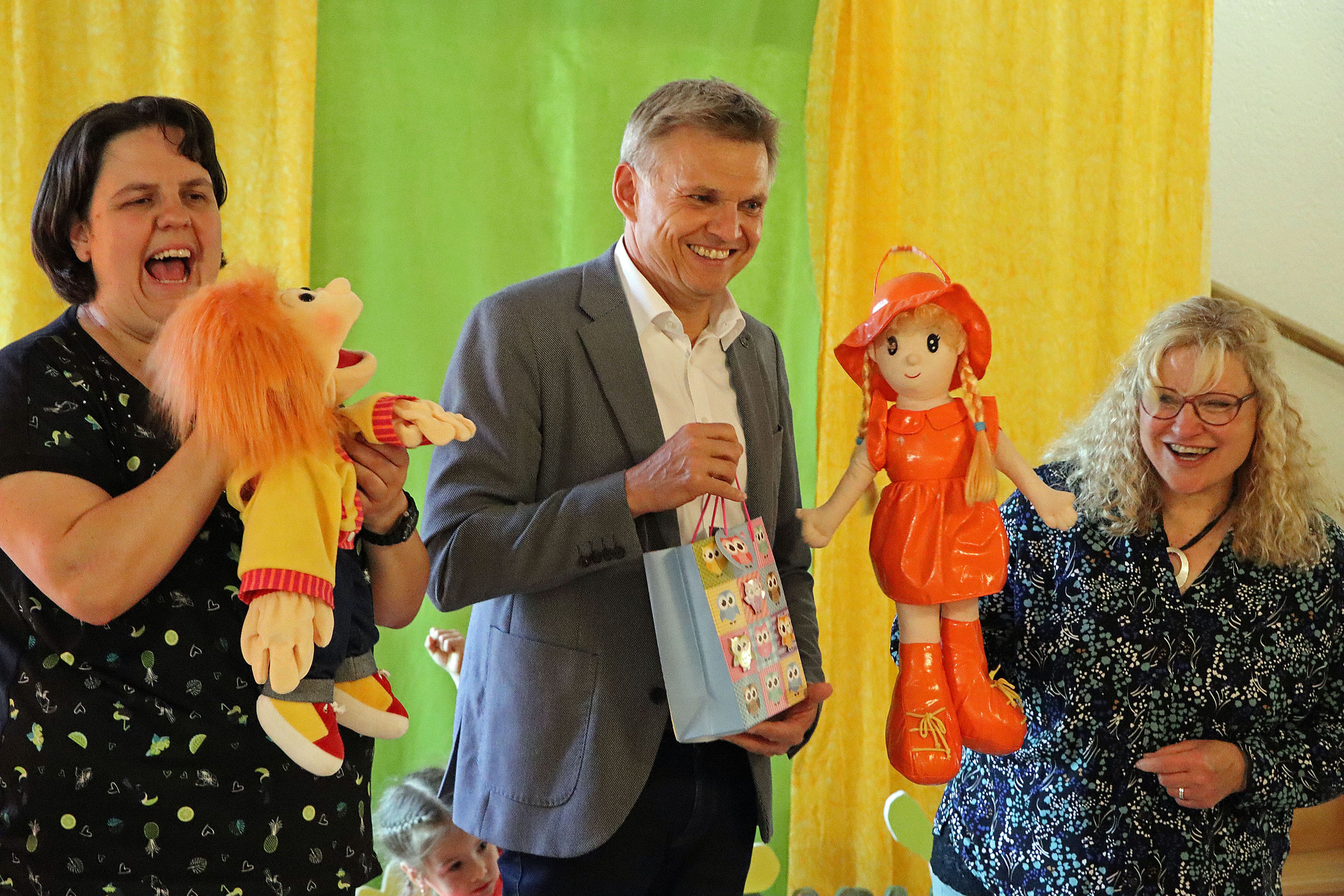 In der Mitte steht ein Mann mit einem Geschenk in der Hand. Es ist der Bürgermeister der Stadt Sonneberg. Links und rechts von ihm steht eine Frau. Sie haben Puppen in der Hand.