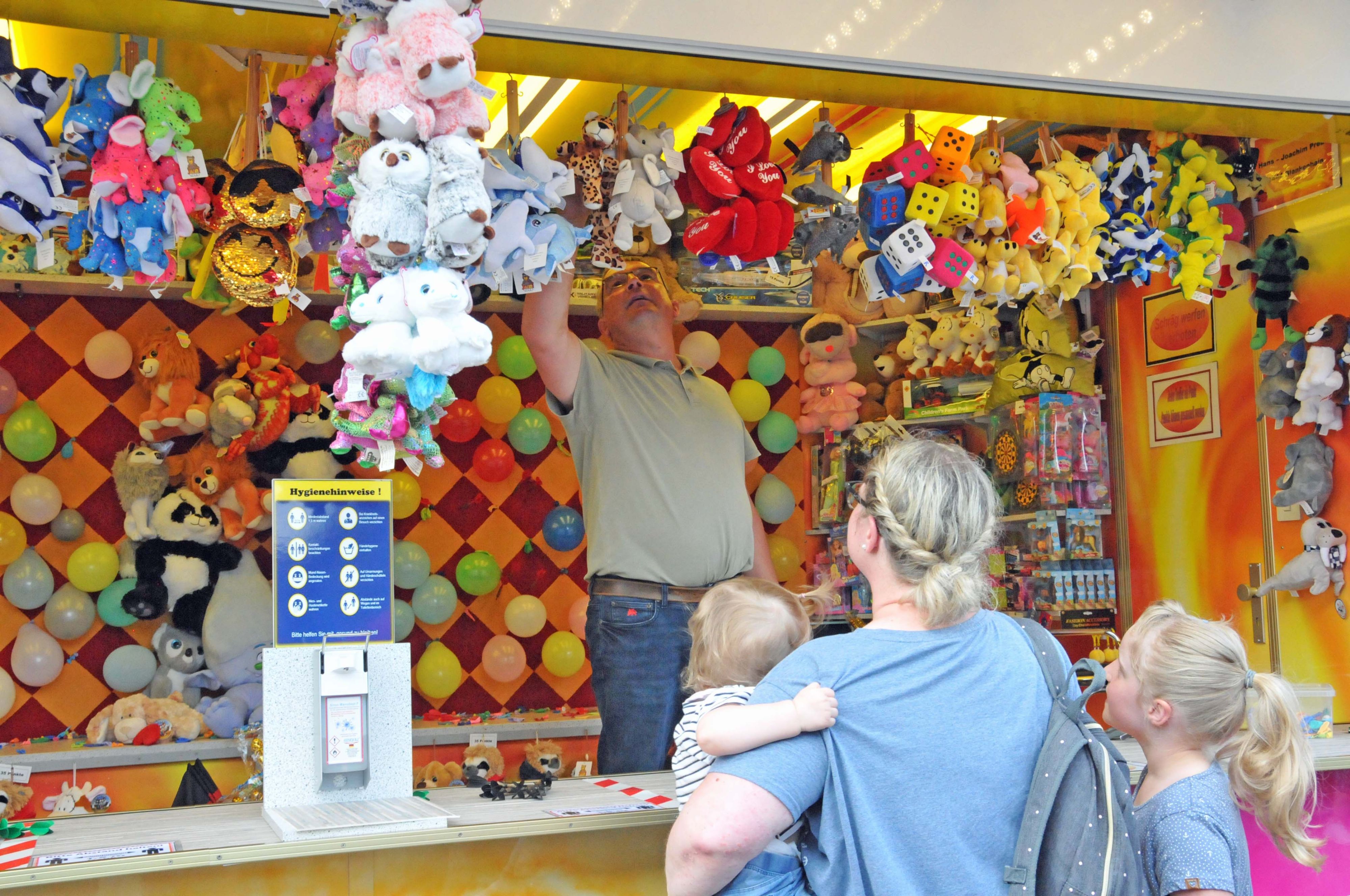 Ein farbenfroher Jahrmarktstand zum Pfeilewerfen. Von der Decke hängen viele Spielsachen.