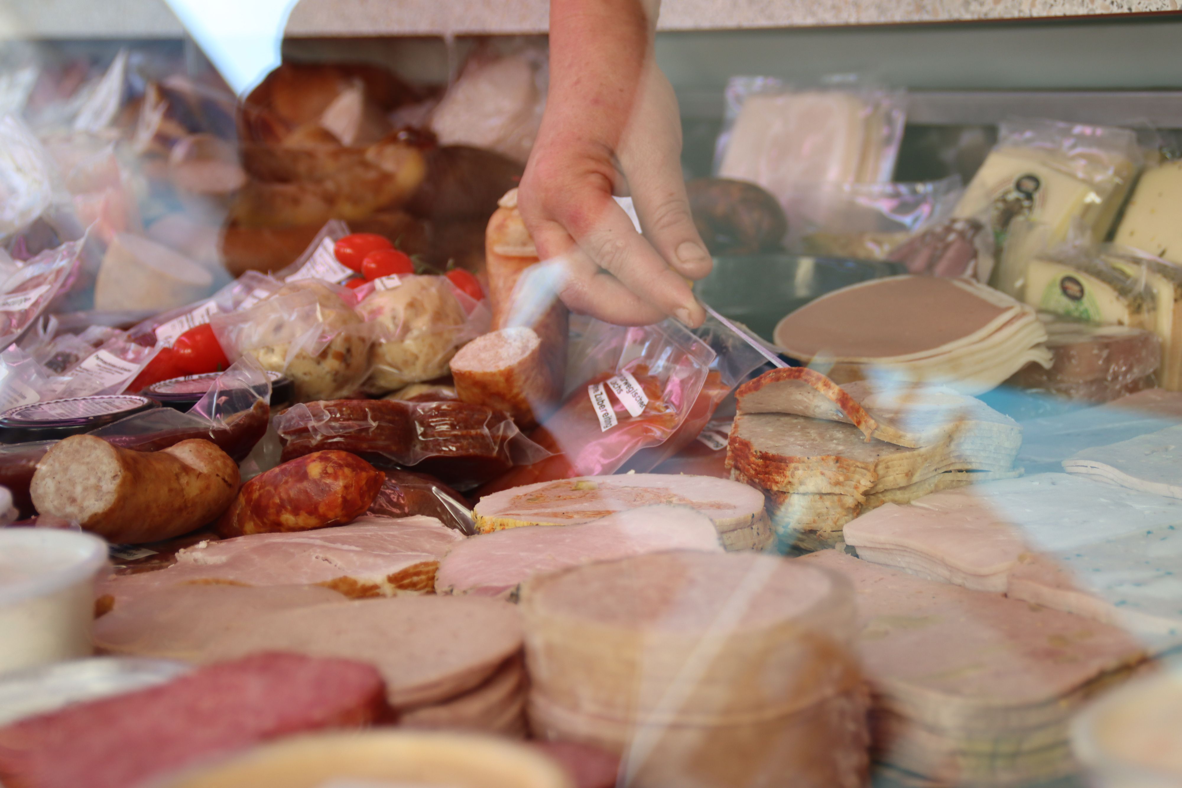 Hinter einer Verkaufstheke liegen Wurst, Fleisch und Käse. Ein Hand greift nach einem abgepacktem Stück Fleisch.