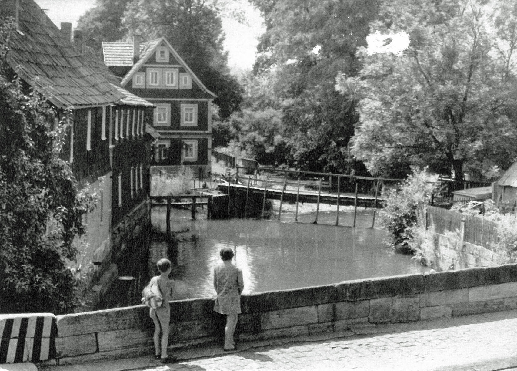Ein Schwarz-Weiß-Foto zeigt zwei Personen, die an einer Brücke stehen und auf einen Fluss schauen. Links stehen alte Häuser.
