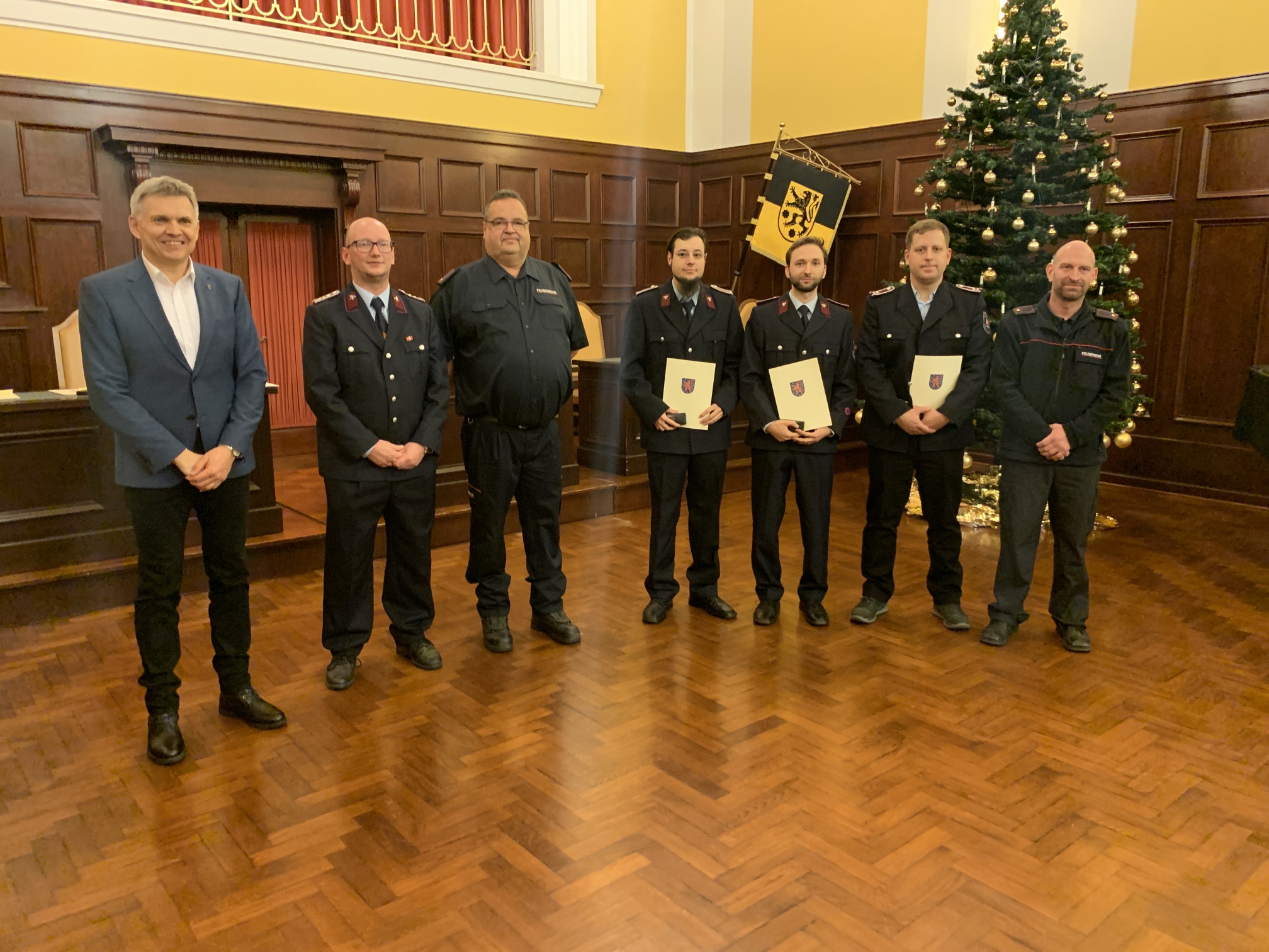 Sechs Männer in Feuerwehr-Uniform stehen in einer Reihe nebeneinander. Drei von ihnen halten eine Urkunde und eine Auszeichnung in den Händen. Links daneben steht der Bürgermeister Dr. Voigt.