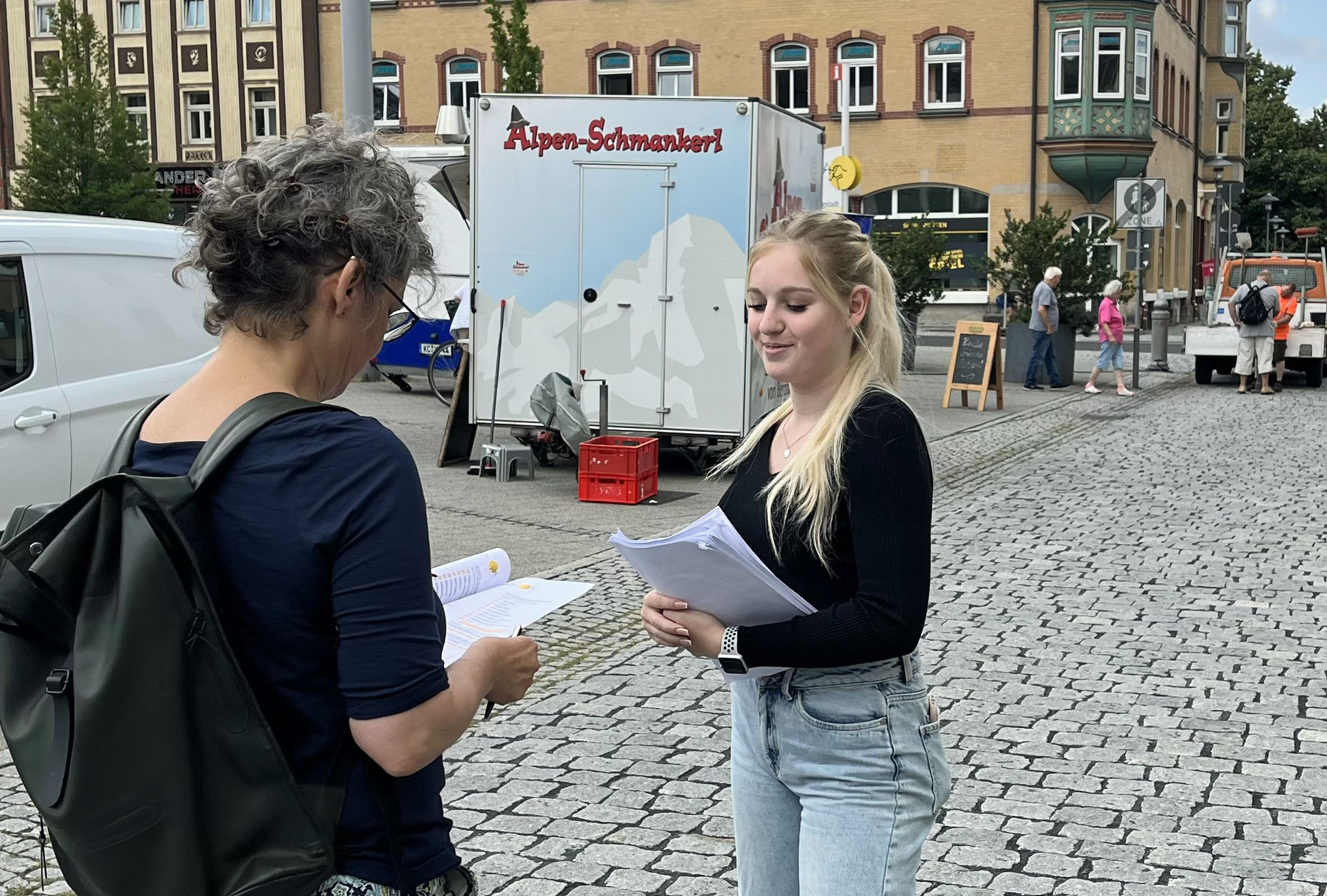 Eine junge Frau gibt einen Fragebogen an eine Besucherin in der Innenstadt.