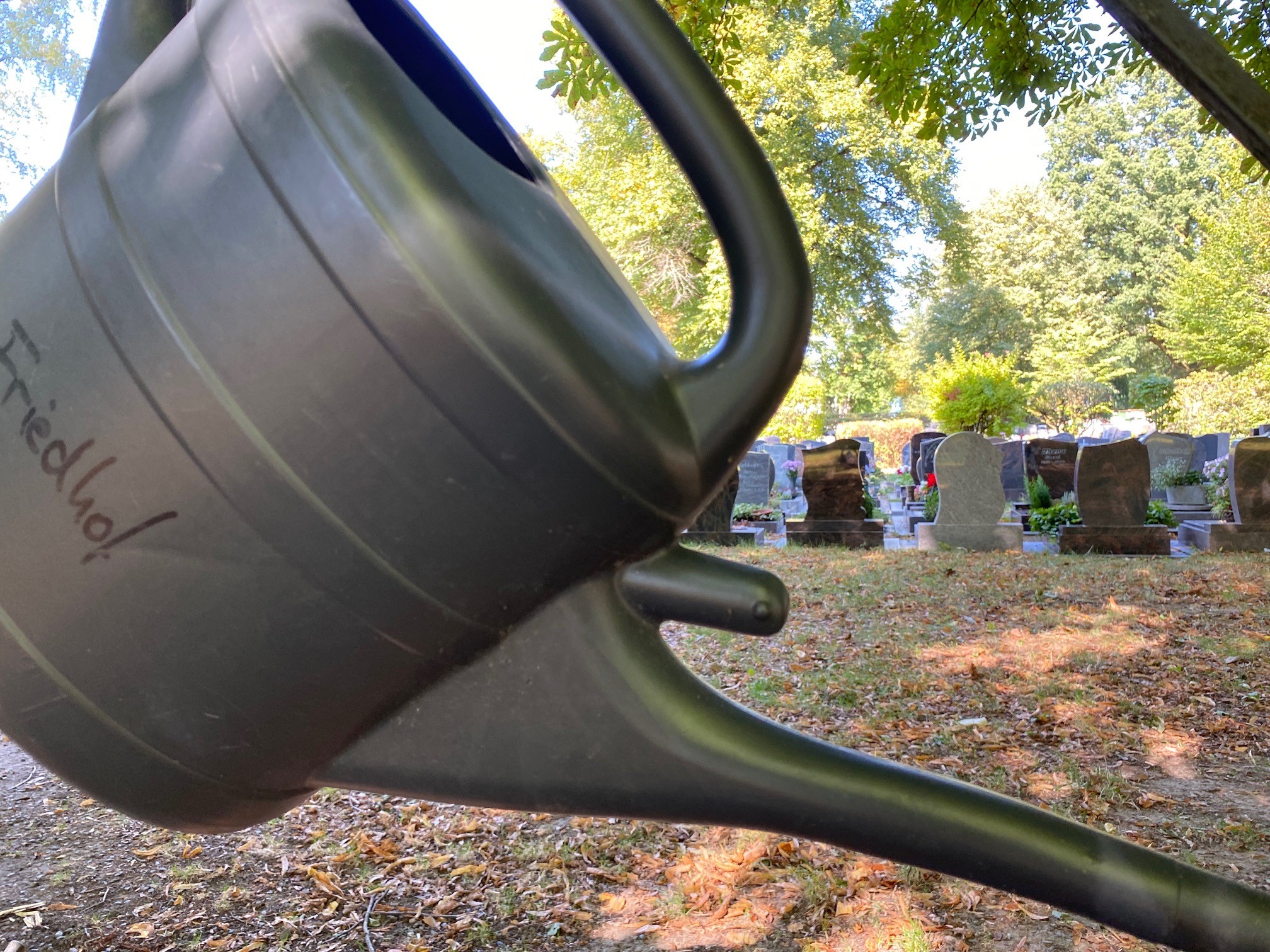 Eine Gießkanne mit der Aufschrift Friedhof hängt an einem Metallgerüst vor einer Grabstelle.