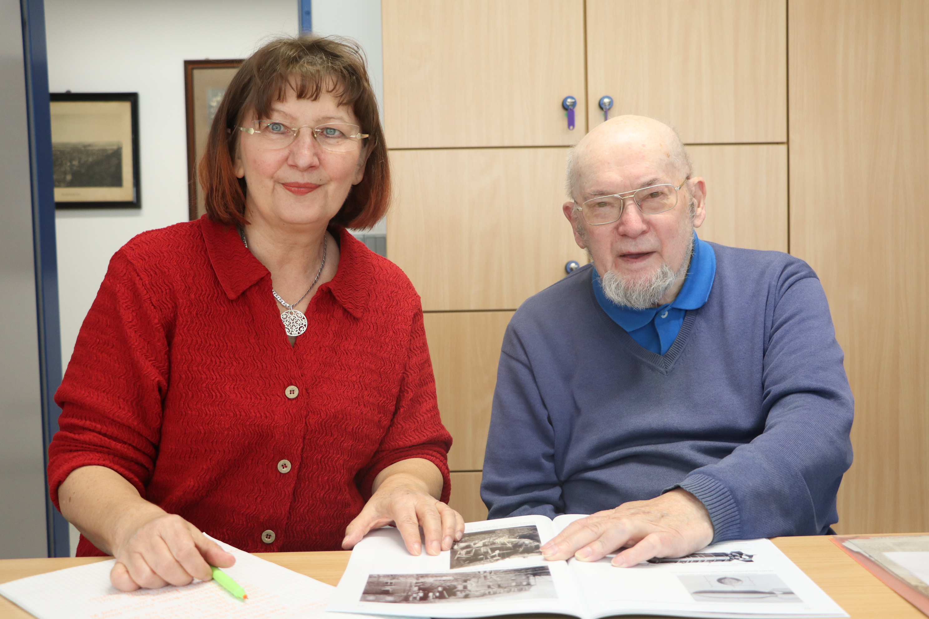 Eine Frau und ein Mann sitzen an einem Schreibtisch und schauen in eine historische Broschüre mit Schwarz-Weiß-Fotos.