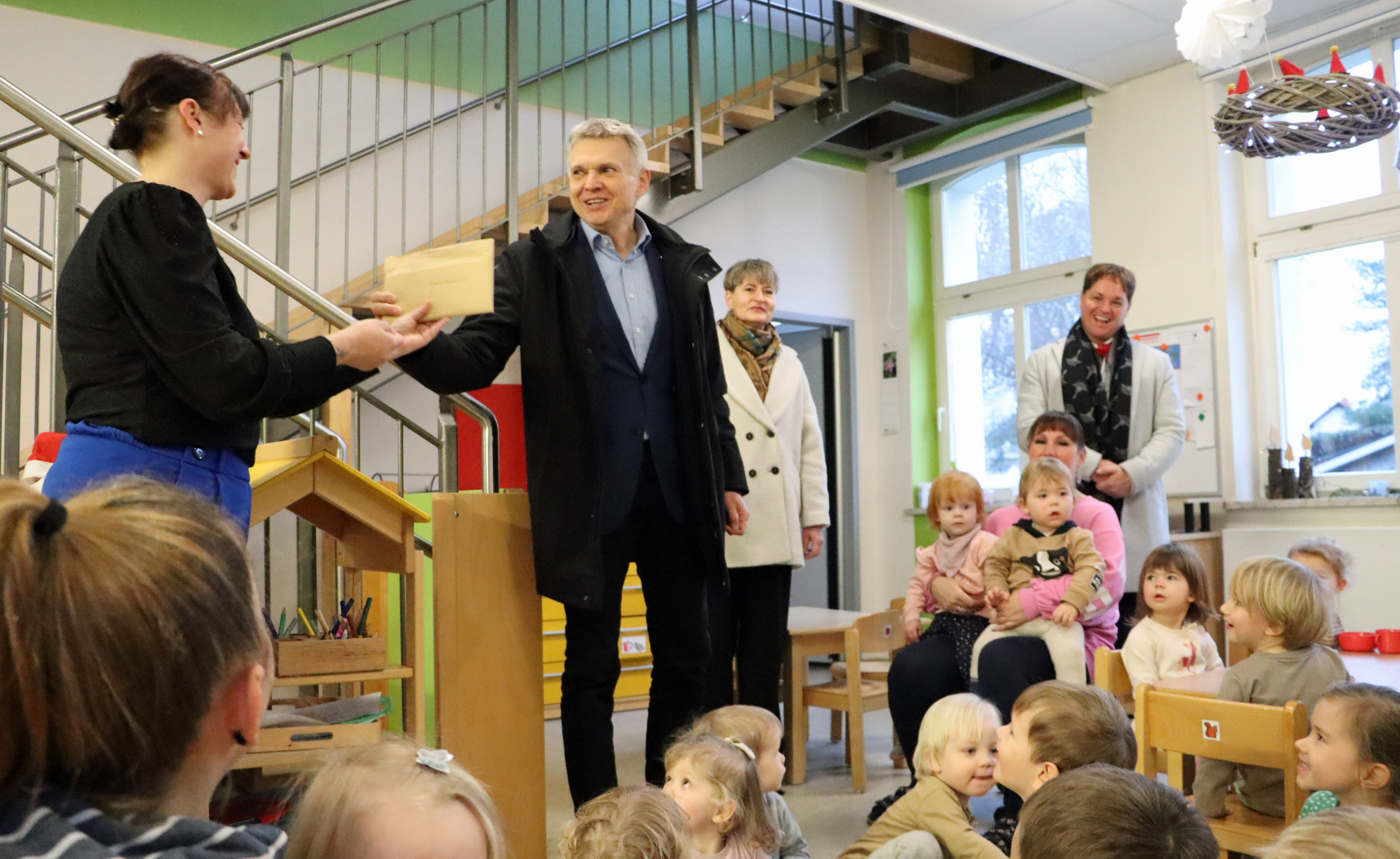 Ein Mann übergibt einen Umschlag an eine Frau in einem Kindergarten. Rundherum sitzen Mädchen und Jungen, die sich über diese Nikolaus-Überraschung freuen.