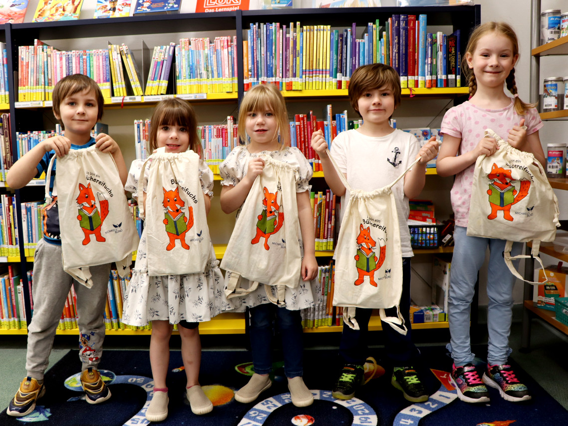 Fünf Vorschüler mit Geschenkrucksäcken vor Bücherregalen in der Bibliothek